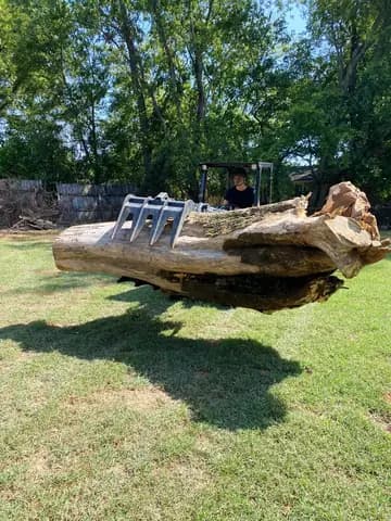 A grapple loader lifts a heavy trunk section off the lawn after a tree removal.